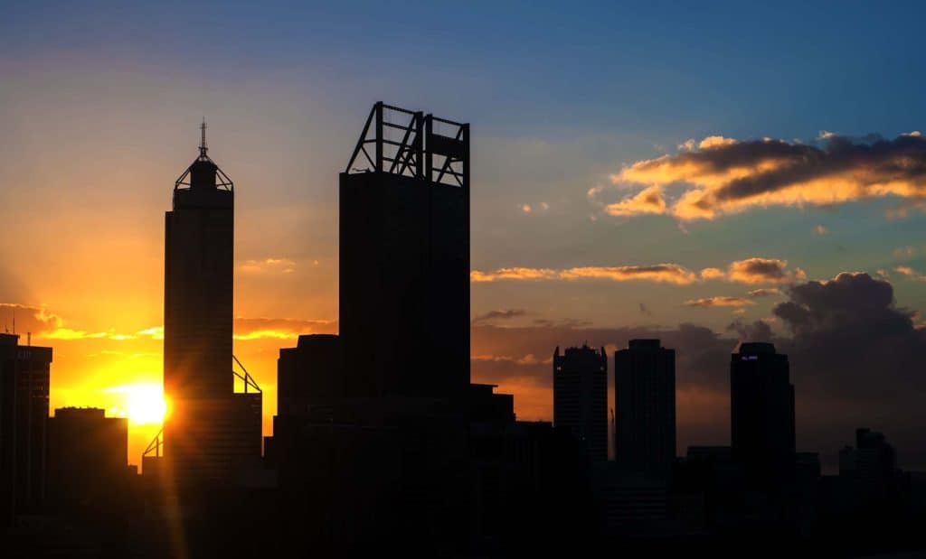 AAB Energy Solar Perth - Silhouette of city skyscrapers at sunset with the sun partially visible behind the buildings and clouds in the sky.