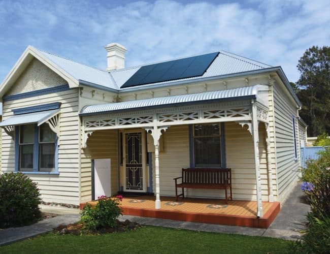 AAB Energy Solar Perth - Single-story cream-colored house with a front porch, wooden bench, and solar panels on the metal roof, surrounded by a lawn and shrubs.